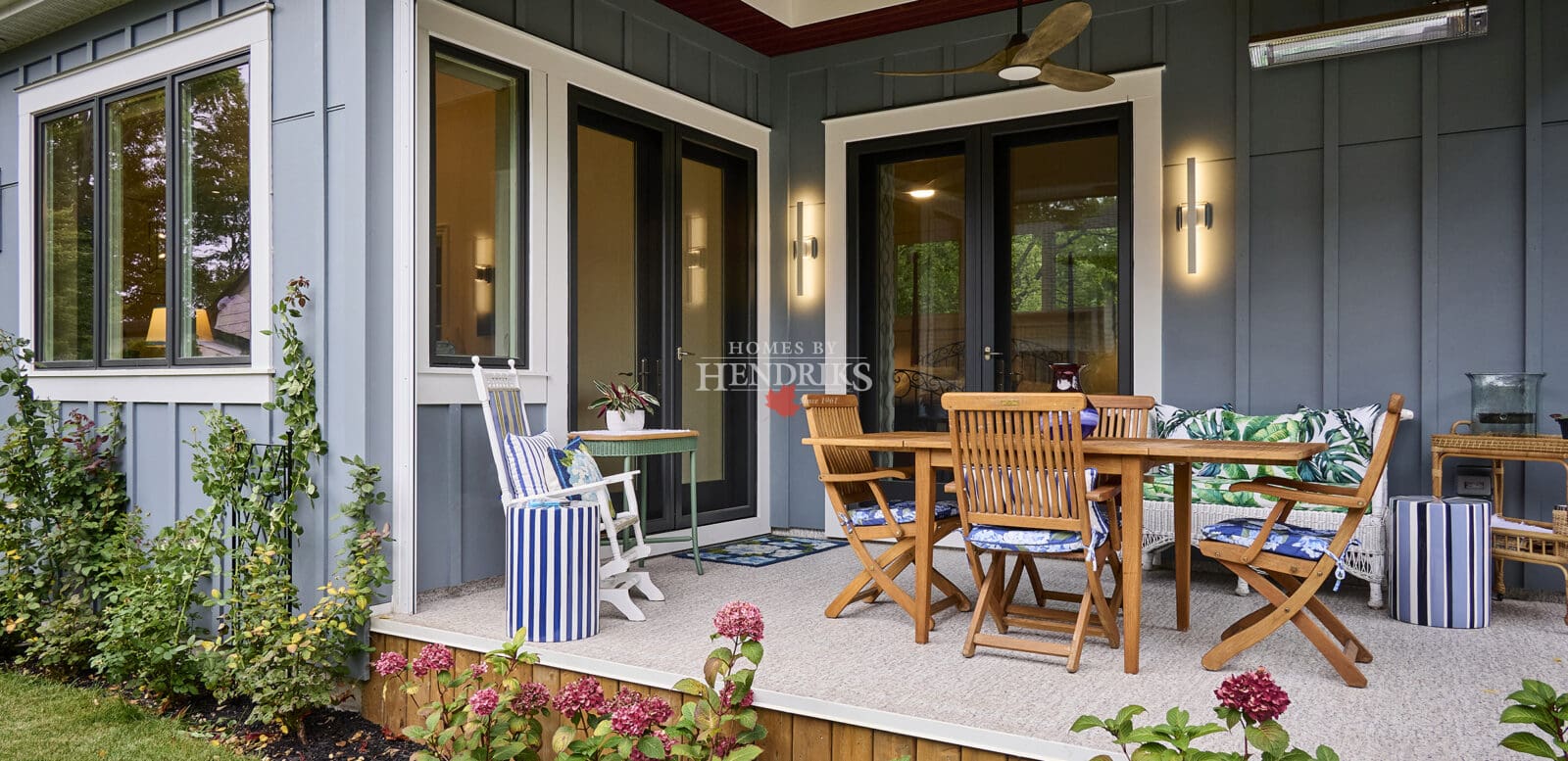 Exterior view of a dining area showcasing teak outdoor furniture and an exterior heater, framed by grey Hardie board and batten siding, emphasizing a comfortable and inviting outdoor setting.