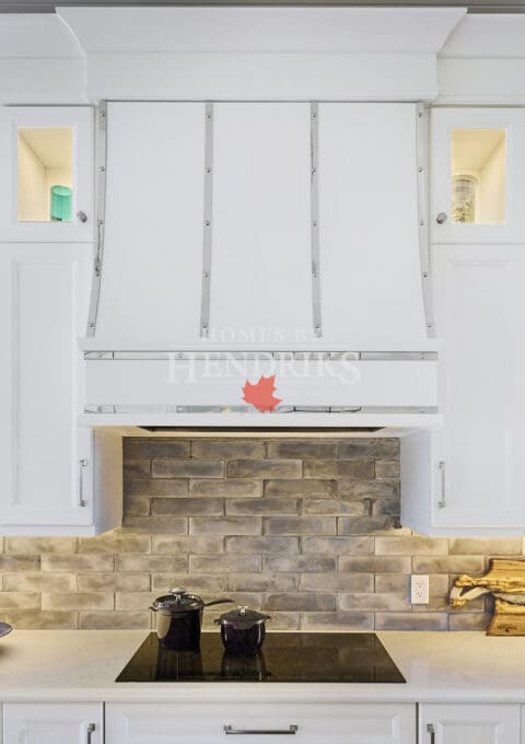 Interior view of a kitchen featuring white cabinetry, a range hood with chrome accents, an induction range, and a stylish brick backsplash, creating a modern and chic cooking space.